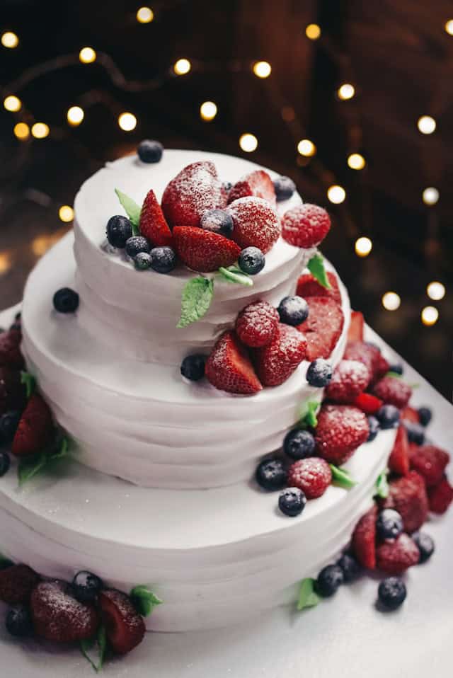 Three-tier white celebration cake decorated with strawberries, blueberries and mint, lightly dusted with sugar, with fairy lights in the background.