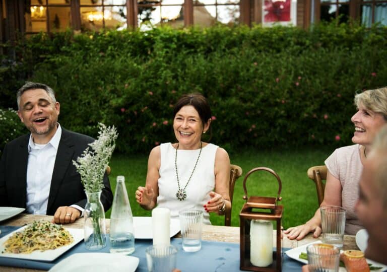 Three adults sit at an outdoor garden table, smiling and chatting as sheet pan ravioli and drinks are served.