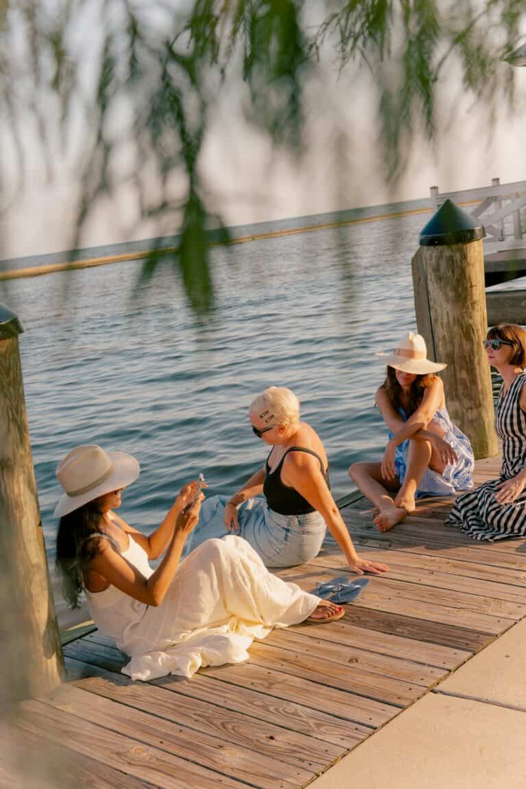 Groups of people relaxing on a wooden dock by the water, wearing sun hats and sunglasses.